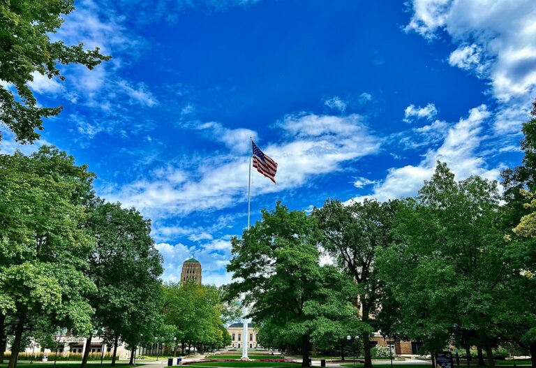 University of Michigan campus with large American flag centered among trees and historic buildings