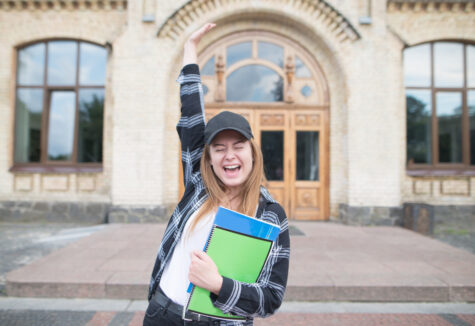 Pretty girl stands on the background of university with books and notebooks in her hands and rejoices