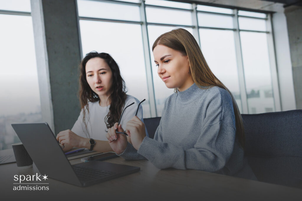 Two women engaged in a coaching session, with one guiding the other who is an intern, seated at a table with an open laptop in front of them displaying a work-related interface.