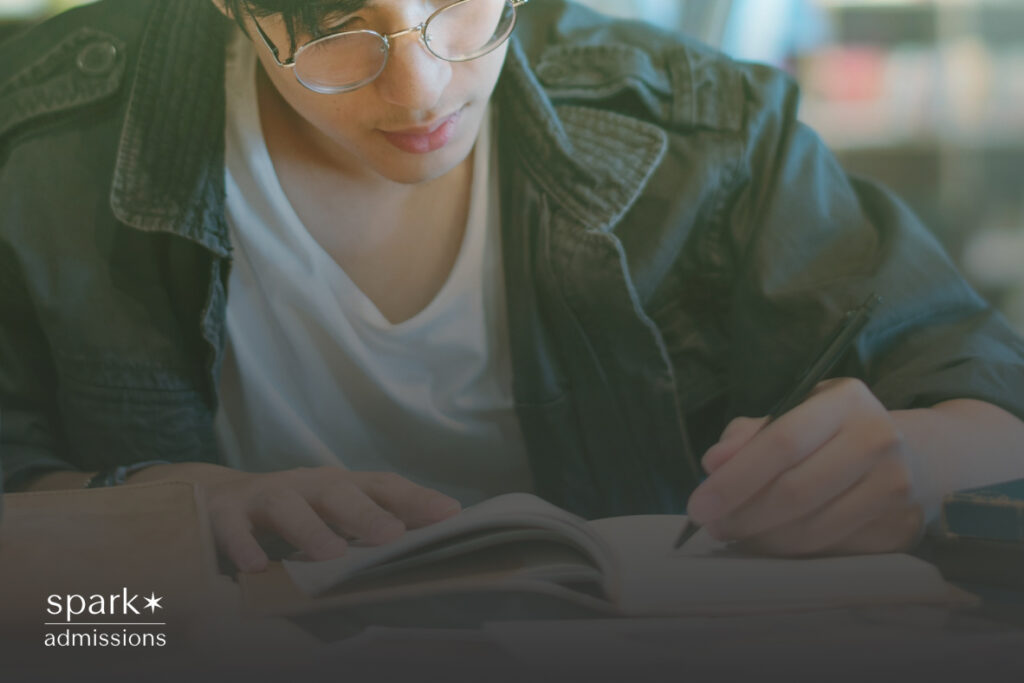 A male student sitting at a desk, studying intently with an open notebook and a pen in hand, surrounded by books and a laptop, in a quiet, focused environment.