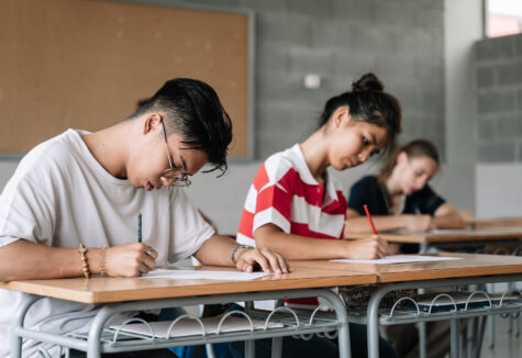 A student sits at a desk in a classroom, focused on writing in a notebook. Other students and a whiteboard with lesson notes are visible in the background.