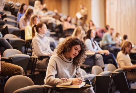 Female college student writing an exam during a class at lecture hall. Her classmate are in the background.