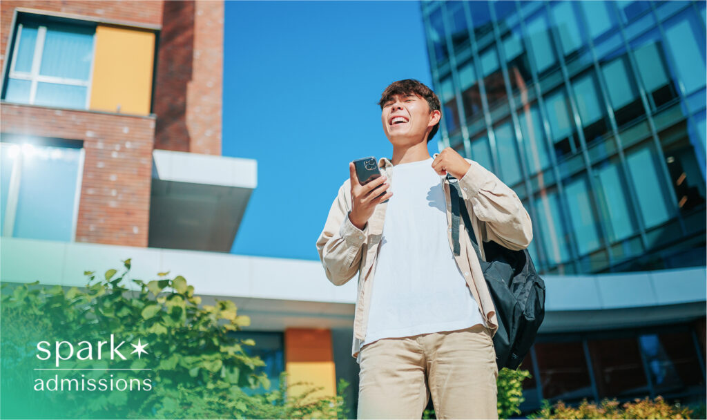 A male student smiling while holding a phone and backpack outside a modern university building